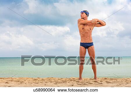 Picture - Muscular male swimmer standing on beach warming up. Fotosearch
