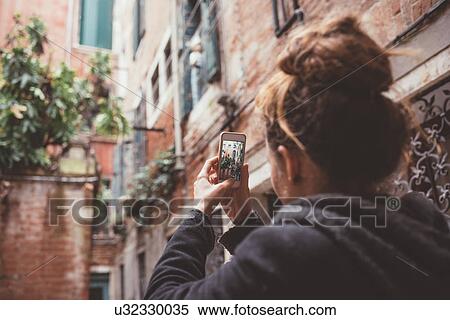 Stock Photography - Over the shoulder view of woman photographing buildings on smartphone, Venice, Italy. Fotosearch