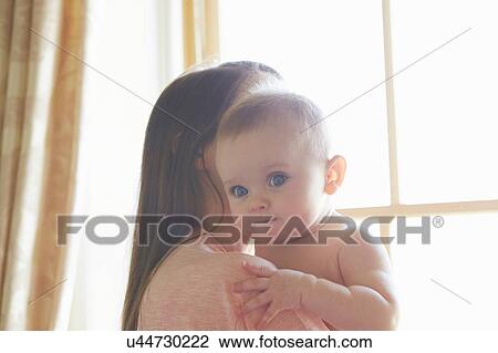 Stock Image - Portrait of baby girl being carried by mother in bedroom. Fotosearch