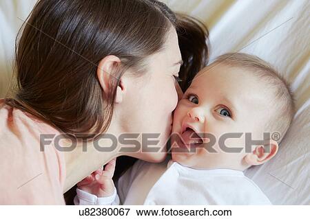 Portrait of cute baby girl being kissed by mother on bed View Large Photo Image Stock Photo - Portrait of cute baby girl being kissed by mother on bed. Fotosearch