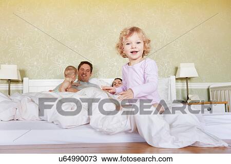 Stock Image - Portrait of cute female toddler sitting on bed with parents and baby sister. Fotosearch