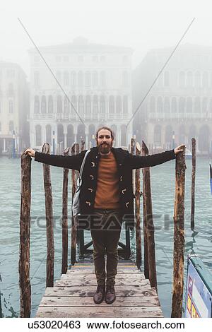 Stock Image - Portrait of mid adult man on misty canal pier, Venice, Italy. Fotosearch