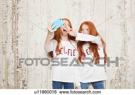 Stock Photograph - Portrait of twin sisters wearing Christmas jumpers, taking selfie using smartphone. Fotosearch