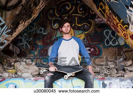 Stock Photography - Portrait of young male skateboarder sitting on graffiti wall at ruined mine. Fotosearch
