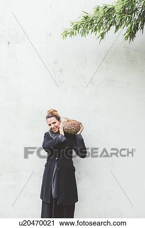 Stock Image - Portrait of young woman carrying pumpkin on shoulder in front of white wall. Fotosearch