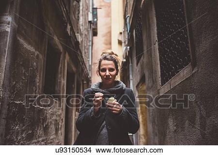Picture - Portrait of young woman eating gelato in dark alley, Venice, Italy. Fotosearch