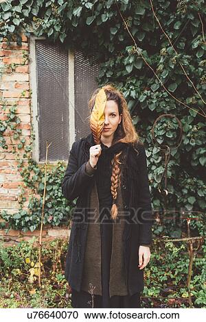 Stock Image - Portrait of young woman holding autumn leaf in front of her face. Fotosearch