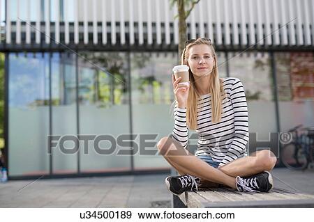 Stock Photo - Portrait of young woman sitting cross legged on city bench with takeaway coffee. Fotosearch