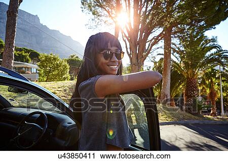 Stock Image - Portrait of young woman standing beside car, leaning on open car door. Fotosearch