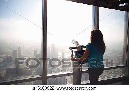 Stock Image - Rear view of female tourist looking through skyscraper window above the city, Dubai, United Arab Emirates. Fotosearch