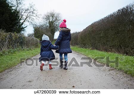 Stock Photo - Rear view of girl and female toddler in knit hats walking along rural road. Fotosearch