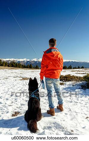 Stock Image - Rear view of man with dog looking out at snow covered landscape. Fotosearch