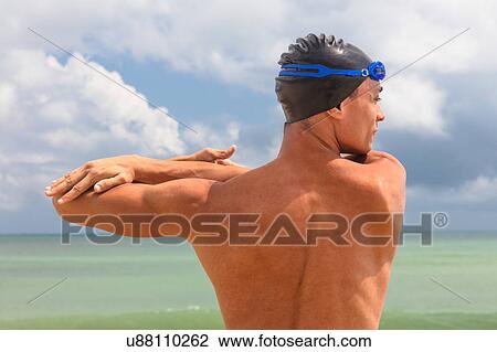 Stock Image - Rear view of muscular male swimmer on beach warming up. Fotosearch