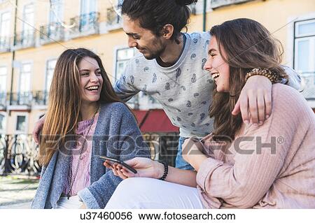 Stock Photograph - Three friends, outdoors, laughing, young woman holding smartphone, Lisbon, Portugal. Fotosearch