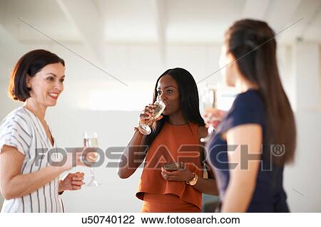 Stock Image - Three women chatting and drinking white wine at art gallery opening. Fotosearch