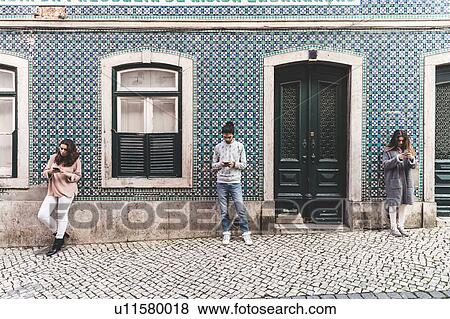 Stock Photo - Three young adults standing in street, looking at their own smartphones, Lisbon, Portugal. Fotosearch
