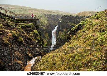 Stock Photo - Tourist on footbridge looking out at canyon waterfall, Fjadrargljufur, Iceland. Fotosearch