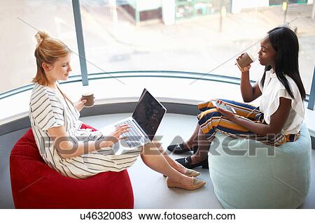 Two businesswomen drinking takeaway coffee sitting on beanbags looking at laptop and smartphone View Large Photo Image Stock Image - Two businesswomen drinking takeaway coffee sitting on beanbags looking at laptop and smartphone. Fotosearch