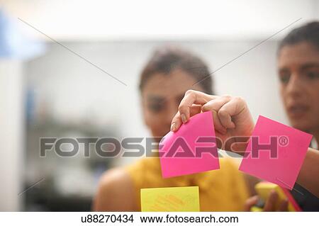 Picture - Two businesswomen removing sticky note from office glass wall. Fotosearch