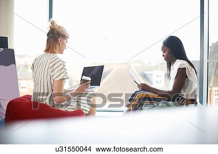 Two businesswomen sitting on beanbags looking at laptop and smartphone View Large Photo Image Picture - Two businesswomen sitting on beanbags looking at laptop and smartphone. Fotosearch