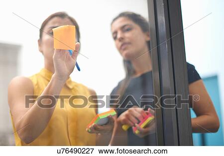 Stock Photo - Two businesswomen sticking sticky note onto office glass wall. Fotosearch