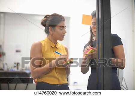 Stock Image - Two businesswomen writing on sticky notes for office glass wall. Fotosearch