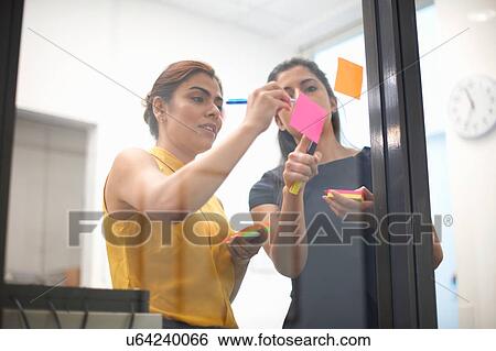 Two businesswomen writing out sticky notes on office glass wall View Large Photo Image Stock Photograph - Two businesswomen writing out sticky notes on office glass wall. Fotosearch
