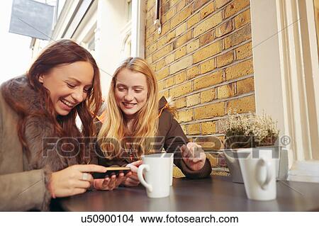 Two female friends, sitting outdoors, having coffee, looking at smartphone View Large Photo Image Picture - Two female friends, sitting outdoors, having coffee, looking at smartphone. Fotosearch