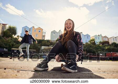 Stock Photo - Two friends fooling around outdoors, young woman sitting on skateboard, laughing, Bristol, UK. Fotosearch