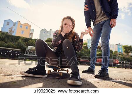 Stock Photography - Two friends fooling around outdoors, young woman sitting on skateboard, laughing, low section, Bristol, UK. Fotosearch