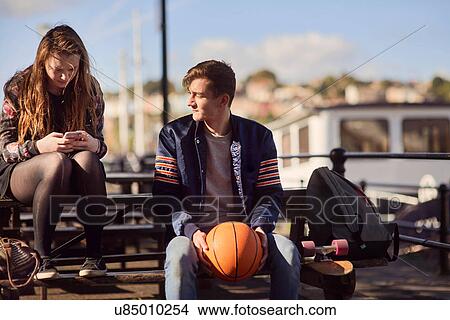 Picture - Two friends sitting outdoors, young man holding basketball, young woman using smartphone, Bristol, UK. Fotosearch