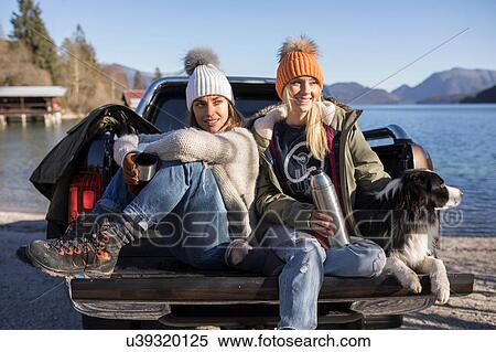 Stock Photography - Two women with coffee and dog sitting on pick up at lakeside in Bavarian Alps. Fotosearch