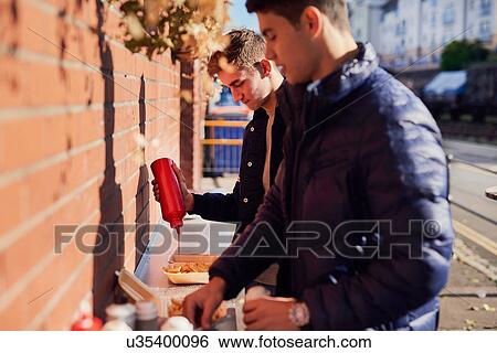 Stock Photograph - Two young men at takeaway food stand, pouring ketchup on food, Bristol, UK. Fotosearch