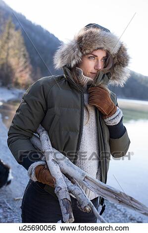 Stock Photograph - Woman in fur trim hood carrying firewood on lakeside in Bavarian Alps. Fotosearch