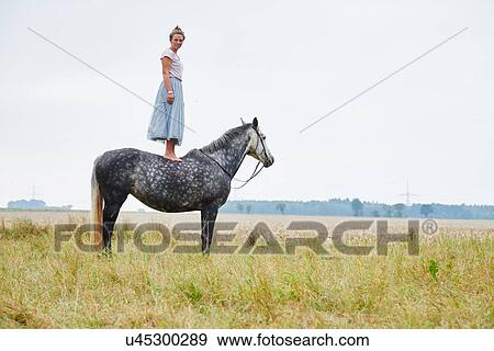 Stock Photo - Woman in skirt standing on top of dapple grey horse in field. Fotosearch