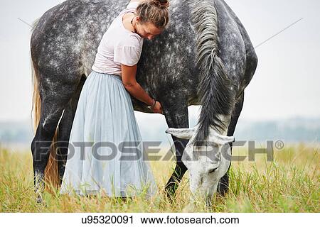 Stock Image - Woman in skirt with arms around dapple grey horse in field. Fotosearch