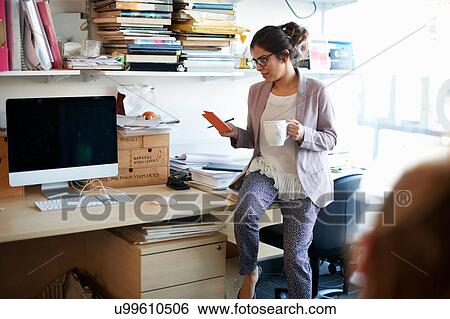 Stock Photograph - Woman sitting on desk looking at notepad. Fotosearch