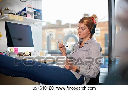Stock Image - Woman wearing headphones with feet up on desk. Fotosearch
