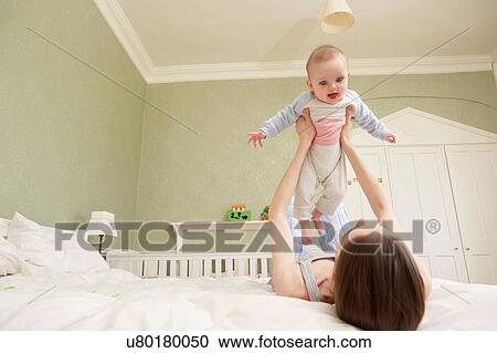 Stock Image - Women lying on bed holding up baby daughter. Fotosearch
