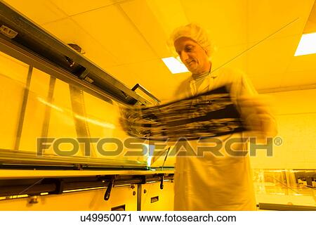 Stock Image - Worker inspecting negative of circuit board in safe yellow light in electronics factory. Fotosearch