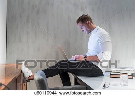 Picture - Young businessman sitting on desk typing on laptop. Fotosearch