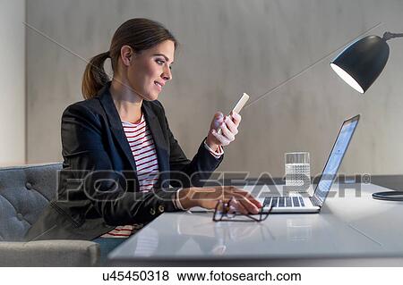 Young businesswoman at desk using laptop and looking at smartphone View Large Photo Image Stock Photo - Young businesswoman at desk using laptop and looking at smartphone. Fotosearch