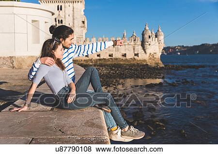 Young couple sitting on wall by sea, Belem Tower in background, looking at view View Large Photo Image Stock Photo - Young couple sitting on wall by sea, Belem Tower in background, looking at view. Fotosearch