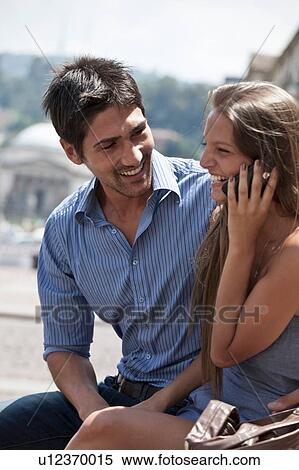 Stock Photography - Young couple sitting outdoors, young woman using smartphone, Turin, Piedmont, Italy. Fotosearch