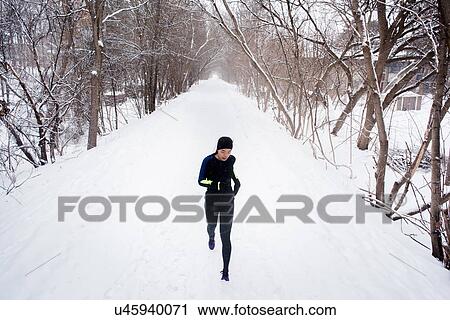 Stock Image - Young female runner in knit hat running in snow covered tree lined park. Fotosearch