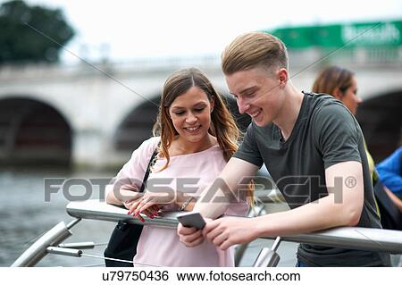 Stock Photograph - Young man and woman beside river, looking at smartphone. Fotosearch