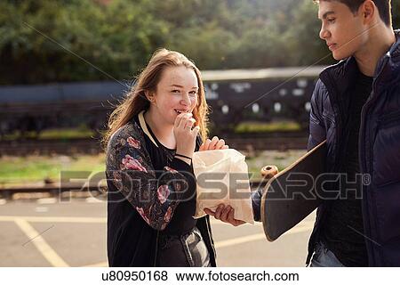 Stock Photo - Young man sharing bag of chips with young woman, skateboard under young man's arm, Bristol, UK. Fotosearch