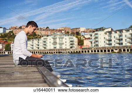 Young man sitting by river, using smartphone, Bristol, UK View Large Photo Image Stock Image - Young man sitting by river, using smartphone, Bristol, UK. Fotosearch