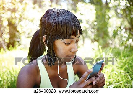 Stock Image - Young woman in rural setting, using smartphone, wearing earphones. Fotosearch