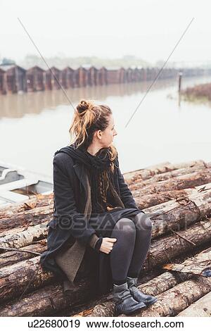 Stock Photo - Young woman sitting on logs on misty canal waterfront. Fotosearch